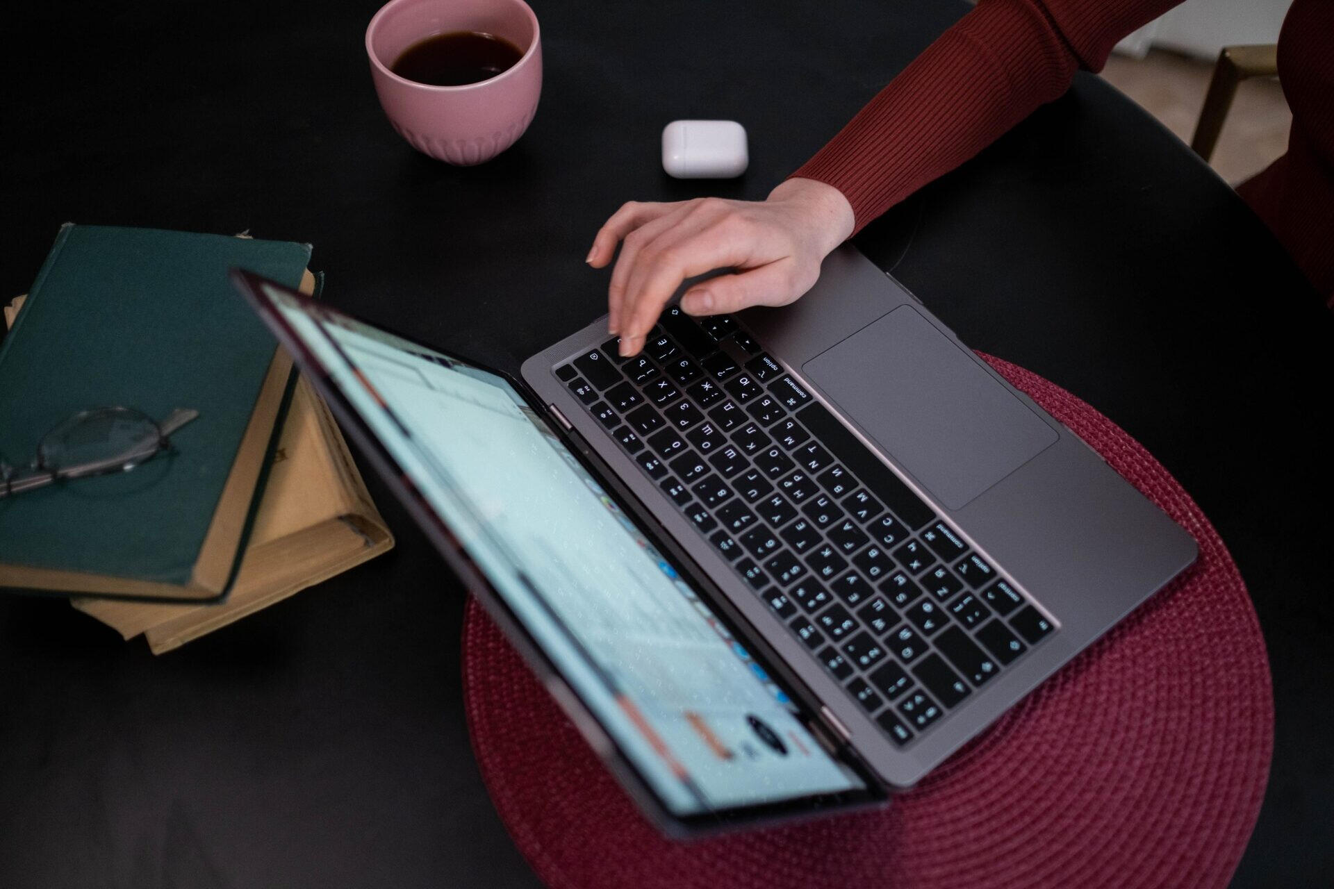Who We Are Hand typing on a laptop beside a stack of notebooks and a coffee cup.